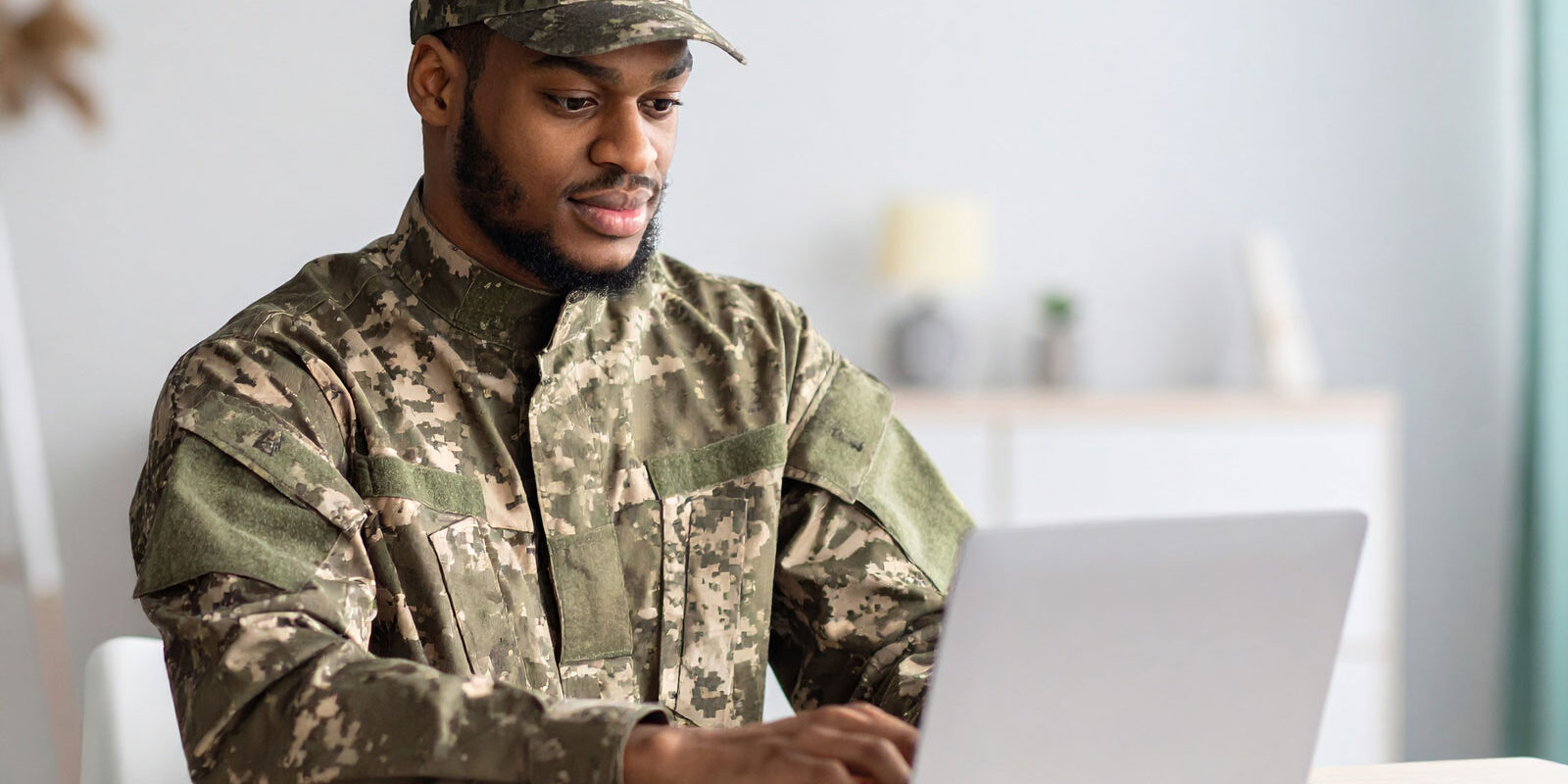 man-on-military-uniform-searching-the-web-on-a-laptop,-seating-on-a-table-at-home
